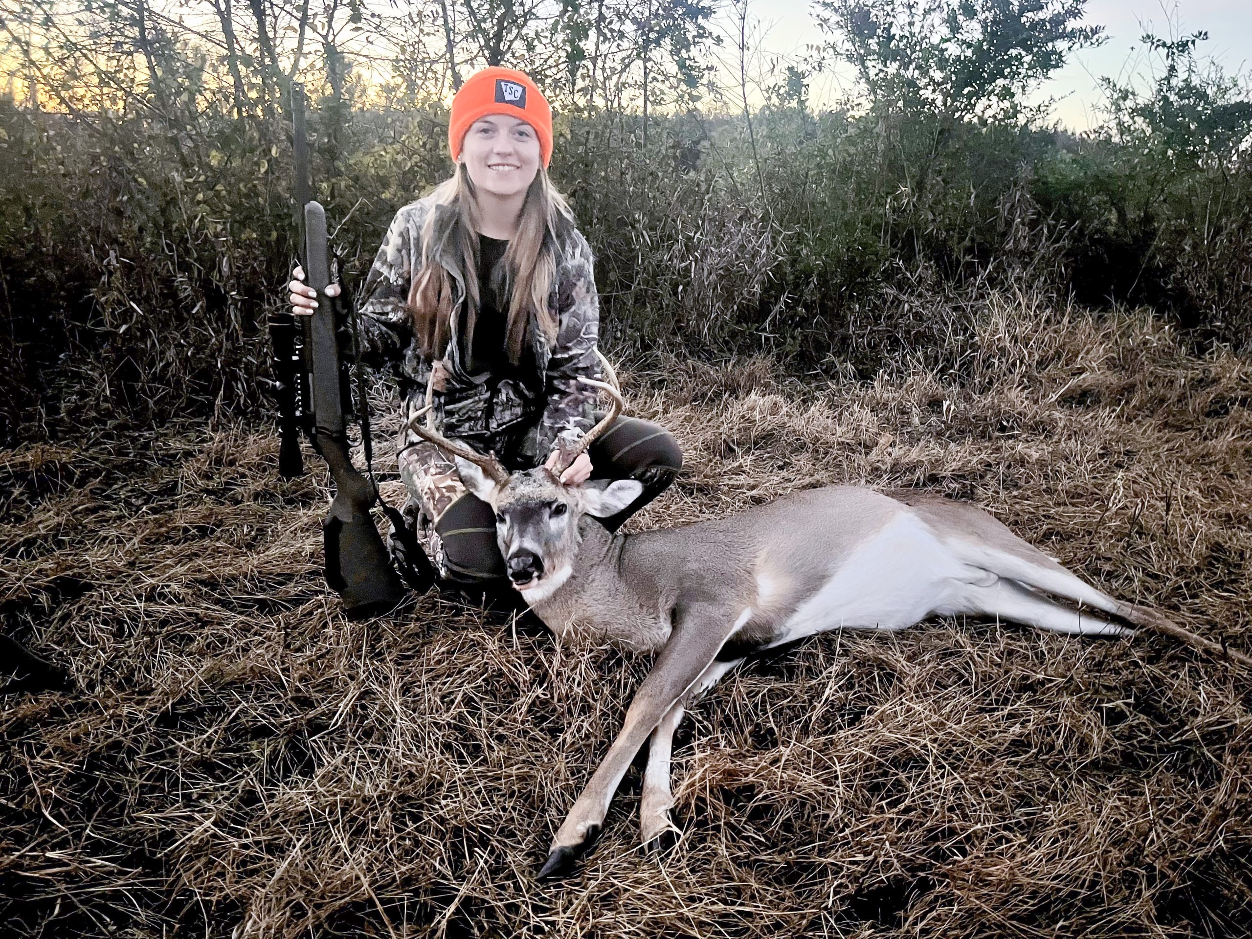 A photo of a young woman holding a gun and kneeling behind a harvested buck deer.