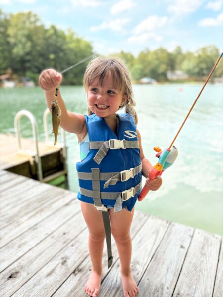 የህይወት ጃኬት የለበሰች ትንሽ ልጅ ዓሣዋን ለካሜራው በገመድ ይዛ ይዛለች።