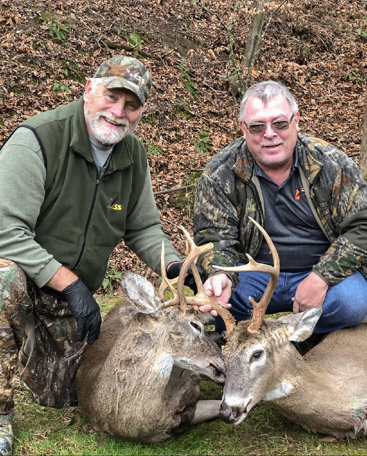 A photo of two men knelling and holding up the antlers of two white-tailed bucks.