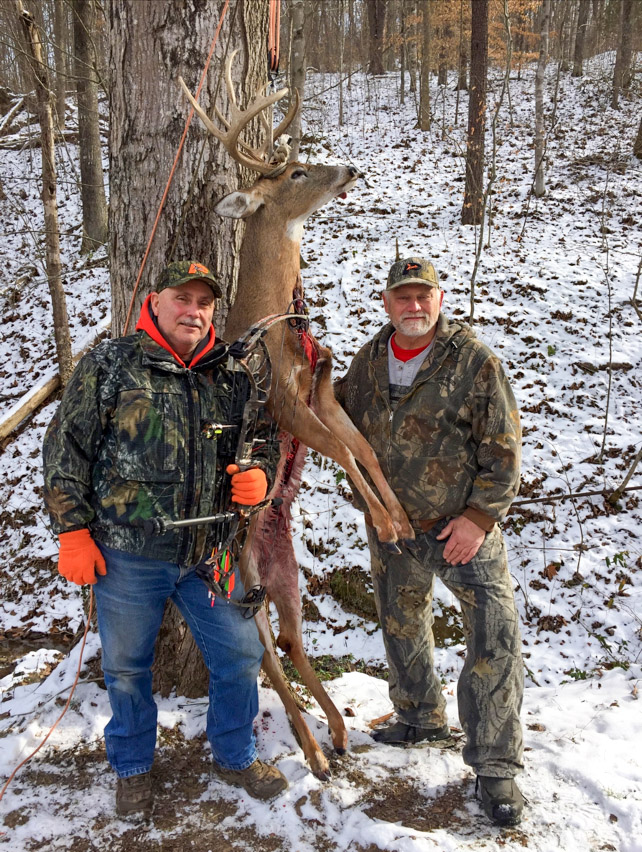 A photo of two men in the woods with snow on the ground, standing by the body of a large white-tailed buck.