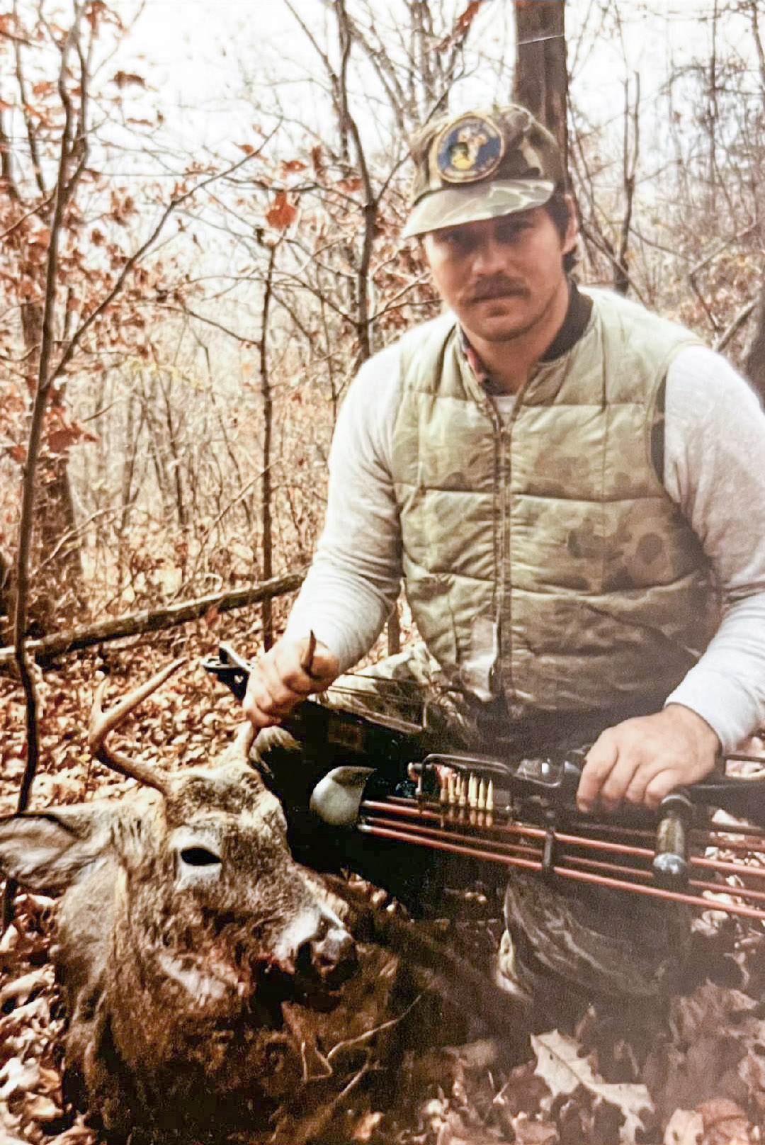 An old photo of a man kneeling in the woods holding a compound bow and the antlers of a white-tailed buck.