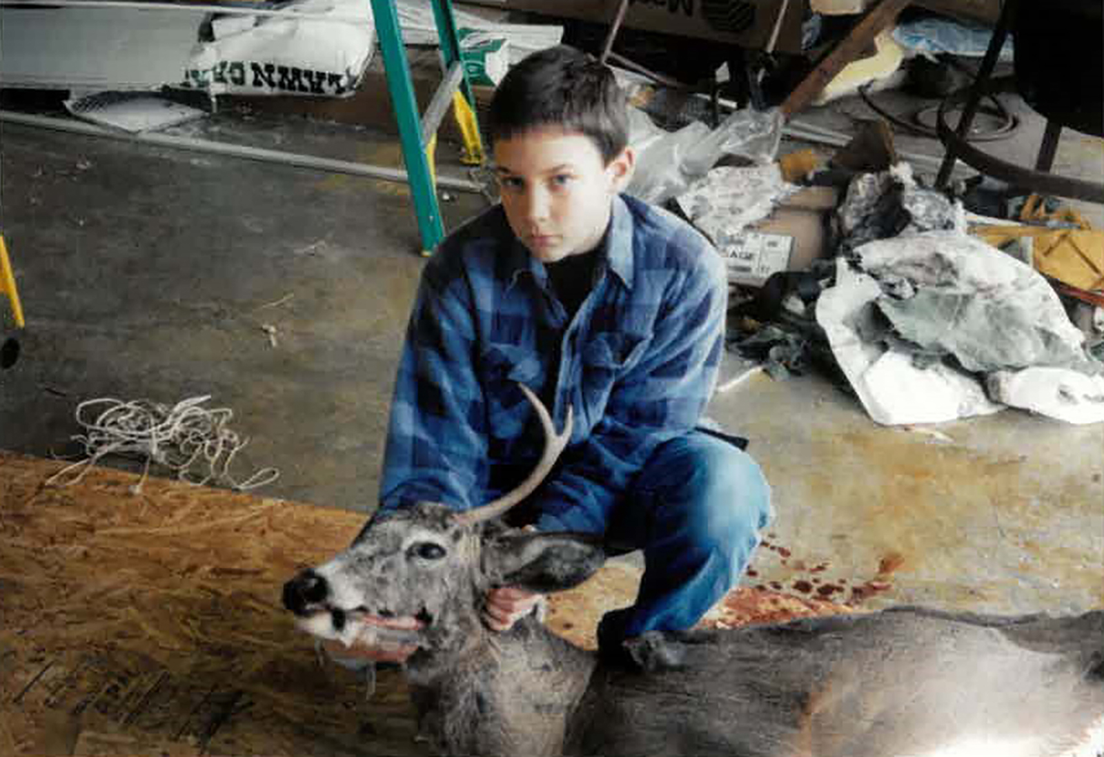 An older photo of a young boy kneeling behind a buck deer in a garage.